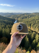 Aus der Ich-Perspektive hält eine Hand eine olivgrüne Kappe mit einem runden "VOGEL-WUID"-Aufnäher in den Vordergrund. Im Hintergrund erstreckt sich eine weite, malerische Landschaft aus dichten Wäldern und einem kleinen Dorf im Tal.