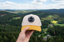 Aus der Ich-Perspektive hält eine Hand eine beige Kappe mit gelbem Schirm und rundem "VOGEL-WUID"-Aufnäher in den Vordergrund. Im Hintergrund erstreckt sich eine weite, malerische Landschaft aus dichten Wäldern und einem kleinen Dorf im Tal, beleuchtet von der Abendsonne.