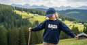 Ein Mädchen in einem dunkelblauen Pullover mit der Aufschrift "BAZI" wandert mit einem Holzstock über eine Almwiese. Mit leicht ausgebreiteten Armen balanciert sie auf dem Hang, während im Hintergrund eine malerische Landschaft aus grünen Hügeln, Holzhütten und einer Bergkette zu sehen ist.