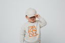Ein kleines Kind in einem hellen Pullover mit orangefarbenem "BAZI"-Aufdruck rückt vor einem neutralen, hellgrauen Studiohintergrund seine helle Kappe zurecht und blickt dabei lächelnd zur Seite.