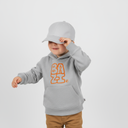 Ein kleiner Junge in einem grauen Kapuzenpullover mit "BAZI"-Aufdruck posiert lächelnd vor einem hellgrauen Studiohintergrund. Er berührt den Schirm seiner grauen Kappe und zieht sie sich spielerisch über die Augen.