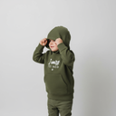 Ein kleines Kind in einem olivgrünen Kapuzenpullover mit der weißen Aufschrift "I wars FEI NED" zieht sich mit beiden Händen die Kapuze tief ins Gesicht. Es blickt zur Seite vor einem neutralen, hellgrauen Studiohintergrund.