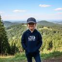 Ein Mädchen in einem dunkelblauen Kapuzenpullover mit der Aufschrift "NIX DERF MA" steht mit den Händen in den Taschen auf einem Aussichtspunkt. Es trägt eine dunkle Kappe, die tief über die Augen gezogen ist, und lächelt sanft. Im Hintergrund erstreckt sich eine weite Landschaft aus bewaldeten Hügeln.