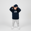 Ein kleines Kind in einem dunkelblauen Kapuzenpullover mit der weißen Aufschrift "NIX DERF MA" zieht sich mit beiden Händen die Kapuze tief ins Gesicht. Es blickt zur Seite vor einem neutralen, hellgrauen Studiohintergrund.