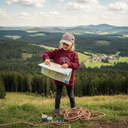 Ein Mädchen in einem weinroten Kapuzenpullover steht auf einem Aussichtspunkt in den Bergen und studiert konzentriert eine große Wanderkarte, die es in den Händen hält. Es zeigt mit dem Finger auf eine Stelle auf der Karte, während zu seinen Füßen ein Seil und Kompasse liegen. Im Hintergrund erstreckt sich eine weite Landschaft aus bewaldeten Hügeln.