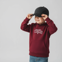 Ein kleiner Junge in einem weinroten Kapuzenpullover mit der Aufschrift "wuide HENA" setzt sich lächelnd mit beiden Händen eine schwarze Kappe auf. Er blickt zur Seite vor einem neutralen, hellgrauen Studiohintergrund.