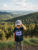 Ein Kind steht auf einer grasbewachsenen Anhöhe und zieht sich mit einer Hand eine graue Kappe über die Augen. Es trägt ein dunkelblaues T-Shirt mit dem lila "GUADE VIBES"-Aufdruck. Im Hintergrund erstreckt sich eine weite Landschaft aus bewaldeten Hügeln.