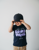 Ein kleiner Junge lächelt, während er sich mit beiden Händen eine schwarze Kappe tief ins Gesicht zieht. Er trägt ein dunkelblaues T-Shirt mit dem großen, lila Aufdruck "i schenk dia GUADE VIBES" vor einem hellgrauen Studiohintergrund.