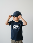 Ein kleiner Junge in einem dunkelblauen T-Shirt mit der weißen Aufschrift "NIX DERF MA" setzt sich lächelnd eine passende Kappe auf. Er blickt zur Seite vor einem neutralen, hellgrauen Studiohintergrund.