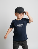 Ein kleiner Junge in einem dunkelblauen T-Shirt mit der weißen Aufschrift "VOGELWUID" berührt den Schirm seiner dunklen Kappe und blickt nachdenklich zur Seite. Er steht vor einem neutralen, hellgrauen Studiohintergrund.