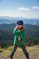Ein Mädchen posiert selbstbewusst mit verschränkten Armen auf einem Aussichtspunkt in den Bergen. Sie trägt einen bunten Strickpullover mit einem Waschbär-Motiv und eine dunkelblaue Kappe mit der Aufschrift "Schlawiner". Im Hintergrund erstreckt sich eine weite Landschaft aus bewaldeten Hügeln.