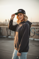 Eine junge Frau mit blonden, gewellten Haaren blickt selbstbewusst über die Schulter in die Kamera, während sie auf einem urbanen Parkdeck steht. Sie trägt einen dunklen Kapuzenpullover und berührt den Schirm ihrer Kappe mit der Aufschrift "#VOGELWUID". Die Szene ist in das warme, goldene Licht des Sonnenuntergangs getaucht.