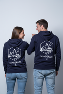 Ein Paar, ein Mann und eine Frau, steht von hinten vor einem hellgrauen Studiohintergrund. Sie tragen identische dunkelblaue Kapuzenpullover, die einen großen, weißen "WANDERLUST"-Aufdruck auf dem Rücken zeigen. Sie blicken sich lächelnd an, während der Mann spielerisch mit dem Daumen auf die Frau zeigt.