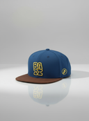 Ein professionelles Produktfoto einer blau-braunen Kappe mit flachem Schirm. Auf der Vorderseite befindet sich der erhabene, gelbe 3D-Schriftzug "BAZI". Die Kappe steht auf einer spiegelnden Oberfläche vor einem hellen Studiohintergrund, was eine deutliche Reflexion erzeugt.