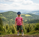 Ein Mädchen in Wanderkleidung, bestehend aus einem pinken T-Shirt mit der Aufschrift "LAUSDEANDL", Shorts und einer Kappe, steht auf einem Aussichtspunkt. Hinter ihr erstreckt sich eine weite Landschaft aus grünen, bewaldeten Hügeln.