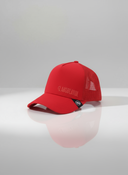 Ein professionelles Produktfoto einer roten Trucker-Kappe. Auf der Vorderseite befindet sich der gestickte Schriftzug "LAUSDEANDL" in einem Ton-in-Ton-Stich. Die Kappe steht auf einer spiegelnden Oberfläche vor einem hellen Studiohintergrund, was eine deutliche Reflexion erzeugt.