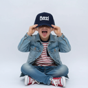 Ein kleiner Junge sitzt im Schneidersitz vor einem hellgrauen Studiohintergrund und macht eine lustige Grimasse. Er trägt eine Jeansjacke und zieht sich mit beiden Händen eine dunkelblaue Kappe mit der weißen Aufschrift "Bazi" über die Augen, während er die Zunge herausstreckt.
