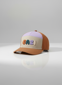 Ein professionelles Produktfoto einer mehrfarbigen Kinder-Kappe im Retro-Stil. Die Kappe kombiniert Farbblöcke in Braun, Lila und Beige und zeigt den bunten, erhabenen 3D-Schriftzug "BAZI" auf der Vorderseite. Sie steht auf einer spiegelnden Oberfläche vor einem hellen Studiohintergrund, was eine deutliche Reflexion erzeugt.