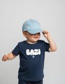 Ein kleiner Junge steht vor einem neutralen, hellgrauen Hintergrund und rückt eine hellblaue Baseball-Kappe zurecht, die seine Augen verdeckt. Er trägt ein dunkelblaues T-Shirt mit dem weißen, aufgeblähten Schriftzug "BAZI".