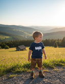 Ein kleiner Junge in traditioneller Lederhose und einem modernen, dunkelblauen T-Shirt mit der Aufschrift "BAZI" steht auf einem Weg am Rande einer blühenden Almwiese. Er blickt in die Ferne auf eine malerische Landschaft aus Hügeln und Bergen, die in das goldene Licht der untergehenden Sonne getaucht ist.