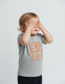 Ein Kleinkind mit blonden Haaren steht vor einem neutralen, hellgrauen Hintergrund und hält sich mit beiden Händen die Augen zu, als würde es Guck-guck spielen. Es trägt ein graues T-Shirt mit dem großen, orangefarbenen Aufdruck "BAZI".