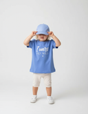 Ein kleines Kind steht vor einem neutralen, hellgrauen Hintergrund und zieht sich mit beiden Händen eine hellblaue Baseball-Kappe über die Augen. Es trägt ein passendes hellblaues T-Shirt mit dem weißen Schriftzug "I wars fei ned".