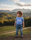 Ein kleines Kind in einem hellblauen T-Shirt mit der Aufschrift "I wars fei ned" steht auf einem Aussichtspunkt in den Bergen. Es blickt nachdenklich zur Seite auf eine weite Landschaft aus bewaldeten Hügeln und einer schroffen Bergkette im Hintergrund.