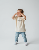 Ein kleines Mädchen mit hochgebundenen Haaren steht lachend vor einem neutralen, hellgrauen Hintergrund und hält sich die Augen mit beiden Händen zu. Es trägt ein cremefarbenes T-Shirt mit dem schwarzen Schriftzug "LAUSDEANDL".