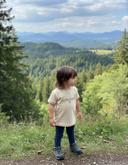 Ein kleines Kind mit langen braunen Haaren steht auf einem Wanderweg und blickt über ein weites Tal mit dichten Wäldern. Am Horizont ist eine blaue Bergkette zu sehen. Das Kind trägt ein cremefarbenes T-Shirt mit dem schwarzen Aufdruck "LAUSDEANDL".