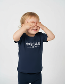 Ein Kleinkind mit blonden Haaren steht lächelnd vor einem neutralen, hellgrauen Hintergrund und verdeckt seine Augen mit beiden Händen. Es trägt ein dunkelblaues T-Shirt mit dem weißen Schriftzug "VOGELWUID".
