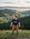 Ein kleiner Junge in einem dunkelblauen T-Shirt mit der Aufschrift "VOGELWUID" steht auf einer grasbewachsenen Anhöhe. Er blickt lächelnd zur Seite, über ein malerisches, nebliges Tal mit einem kleinen Dorf, dichten Wäldern und Bergen in der Ferne.