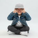 Ein Junge in einer Jeansjacke sitzt im Schneidersitz vor einem hellgrauen Studiohintergrund. Er trägt eine graue Kappe mit dem weißen Schriftzug "Bazi" und hält sich spielerisch die Hände vor die Augen.