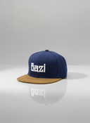 Ein professionelles Produktfoto einer blau-braunen Kappe mit flachem Schirm. Auf der Vorderseite befindet sich der erhabene, weiße 3D-Schriftzug "Bazi". Die Kappe steht auf einer spiegelnden Oberfläche vor einem hellen Studiohintergrund und erzeugt eine deutliche Reflexion.