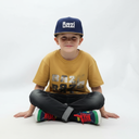 Ein Junge mit Sommersprossen sitzt im Schneidersitz vor einem weißen Studiohintergrund und lächelt freundlich in die Kamera. Er trägt eine dunkelblaue Kappe mit braunem Lederschirm und dem weißen 3D-Schriftzug "Bazi".