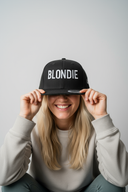 Eine lächelnde Frau mit langen blonden Haaren sitzt vor einem hellgrauen Studiohintergrund und zieht sich mit beiden Händen eine schwarze Kappe mit dem weißen Schriftzug "BLONDIE" tief ins Gesicht, sodass ihre Augen verdeckt sind, aber ihr breites Lächeln sichtbar ist.