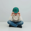 Ein kleiner Junge sitzt im Schneidersitz vor einem hellgrauen Studiohintergrund und hält sich mit beiden Händen eine grüne Trucker-Kappe mit einem gestickten "Bulldog"-Traktor-Motiv vor das Gesicht.