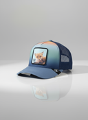 Ein professionelles Produktfoto einer mehrfarbigen Trucker-Kappe. Die Vorderseite hat einen Farbverlauf von Blau zu Orange und im Zentrum befindet sich ein quadratischer Aufnäher, der einen Fuchswelpen und den Schriftzug "FUX" zeigt. Die Kappe steht auf einer spiegelnden Oberfläche vor einem hellen Studiohintergrund, was eine deutliche Reflexion erzeugt.