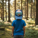 Ein kleiner Junge, von hinten fotografiert, steht in einem sonnendurchfluteten Wald. Er trägt ein blaues T-Shirt und eine mehrfarbige Kappe mit einem Fuchs-Aufnäher auf der Vorderseite.