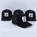 Ein Produktfoto von vier identischen schwarzen Trucker-Kappen, die auf einer weißen Fläche arrangiert sind. Jede Kappe hat einen quadratischen Aufnäher auf der Vorderseite, der eine Ziege und den Schriftzug "GOASS" zeigt. Eine Kappe ist im scharfen Vordergrund, die anderen drei sind dahinter leicht unscharf.