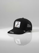 Ein professionelles Produktfoto einer schwarzen Trucker-Kappe. Auf der Vorderseite befindet sich ein quadratischer Aufnäher, der eine Ziege und den Schriftzug "GOASS" zeigt. Die Kappe steht auf einer spiegelnden Oberfläche vor einem hellen Studiohintergrund, was eine deutliche Reflexion erzeugt.