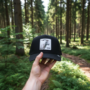 Aus der Ich-Perspektive hält eine Hand eine schwarze Kappe mit einem Ziegen-Aufnäher in den Vordergrund. Im Hintergrund erstreckt sich ein sonnendurchfluteter Waldweg, der von hohen Nadelbäumen und grünem Unterholz gesäumt ist.