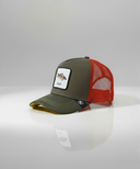Ein professionelles Produktfoto einer Trucker-Kappe. Die Kappe hat eine olivgrüne Vorderseite und einen roten Mesh-Einsatz. Im Zentrum befindet sich ein quadratischer Aufnäher, der einen Karpfen und den Schriftzug "Karpfa" zeigt. Die Kappe steht auf einer spiegelnden Oberfläche vor einem hellen Studiohintergrund und erzeugt eine deutliche Reflexion.