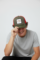 Ein Mann mit einem breiten Lächeln sitzt vor einem hellgrauen Studiohintergrund und zieht sich mit einer Hand spielerisch seine Kappe ins Gesicht. Die Kappe ist olivgrün sowie rot und hat einen Aufnäher mit einem Karpfen und dem Schriftzug "Karpfa".