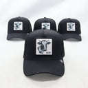 Ein Produktfoto von vier identischen schwarzen Trucker-Kappen, die auf einer weißen Fläche arrangiert sind. Jede Kappe hat einen quadratischen Aufnäher auf der Vorderseite, der einen Kuhkopf und den Schriftzug "KUAH" zeigt. Eine Kappe ist im scharfen Vordergrund, die anderen drei sind dahinter leicht unscharf.