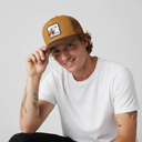 Ein Mann mit einem sympathischen Lächeln und Tattoos auf den Armen sitzt vor einem hellgrauen Studiohintergrund. Er berührt leicht den Schirm seiner braunen Kappe, die einen Aufnäher mit einer Kuh und dem Schriftzug "KUAH" trägt.