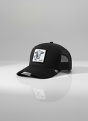Ein professionelles Produktfoto einer schwarzen Trucker-Kappe. Auf der Vorderseite befindet sich ein quadratischer Aufnäher, der einen Kuhkopf und den Schriftzug "KUAH" zeigt. Die Kappe steht auf einer spiegelnden Oberfläche vor einem hellen Studiohintergrund, was eine deutliche Reflexion erzeugt.