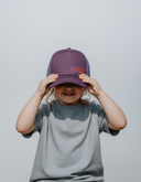 Ein lächelndes Kind in einem grauen T-Shirt hält sich mit beiden Händen eine lila Trucker-Kappe vor das Gesicht. Die Kappe hat den roten Schriftzug "LAUSDEANDL" auf der Vorderseite.