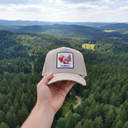 Aus der Ich-Perspektive hält eine Hand eine beige Kappe mit einem Eichhörnchen-Aufnäher in den Vordergrund. Im Hintergrund erstreckt sich eine weite, malerische Landschaft aus dichten Wäldern und sanften Hügeln unter einem leicht bewölkten Himmel.