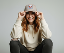 Eine Frau mit einem breiten Lächeln sitzt vor einem hellgrauen Studiohintergrund und hält mit beiden Händen eine beige Kappe fest. Auf der Kappe befindet sich ein Aufnäher mit der Abbildung eines Eichhörnchens und dem Schriftzug "OACHKATZL".