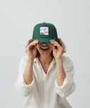 Eine Frau lacht ausgelassen, während sie vor einem hellgrauen Studiohintergrund eine grüne Kappe mit beiden Händen über ihre Augen zieht. Auf der Kappe ist ein Aufnäher mit einem Schaf und dem Schriftzug "SCHOOF" zu sehen.